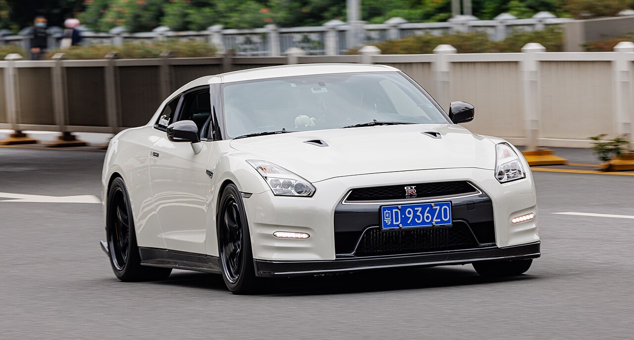 A white Nissan GT-R R35 is captured in motion on a city road. The car features aggressive styling, black wheels, LED headlights, hood vents, and a front grille with the GT-R badge. A blue license plate is visible, and the background includes railings and greenery