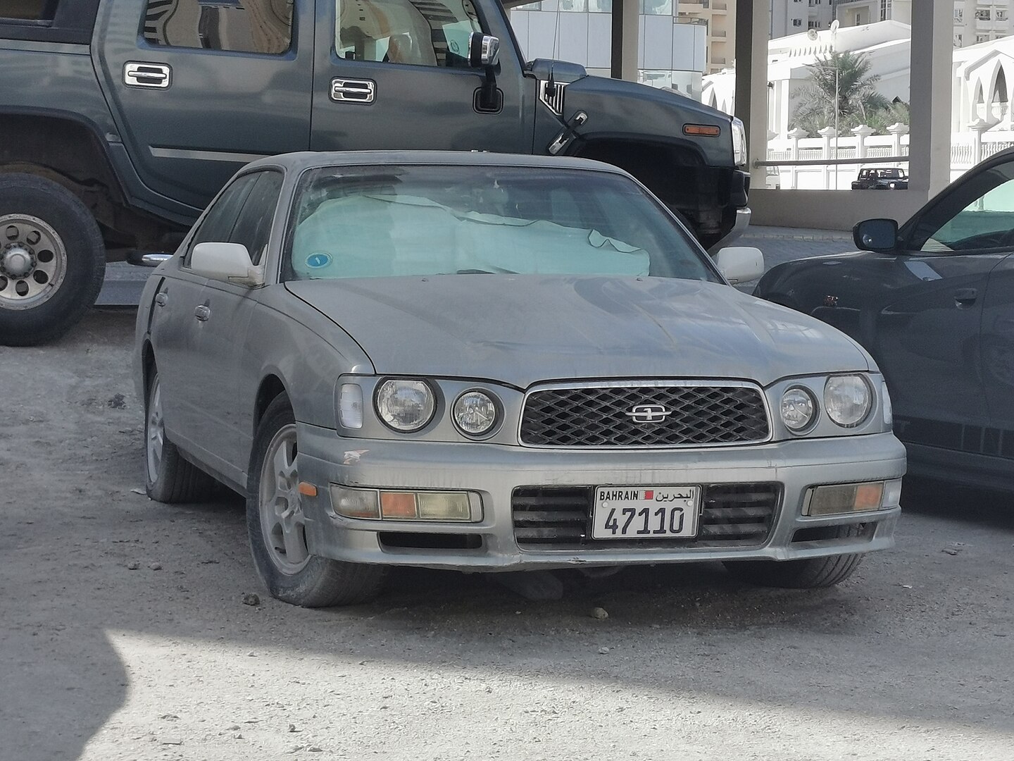 A dusty silver Nissan Gloria sedan with a Bahrain license plate is parked on a gravel surface. The car has round headlights, a mesh grille, and visible wear on the bumper. Other vehicles, including a black Hummer, are parked nearby under a shaded area