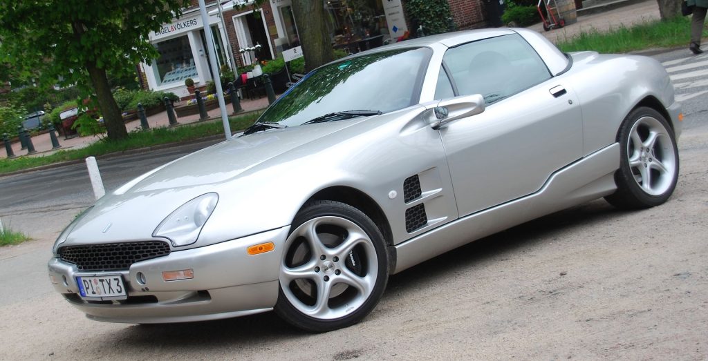 Silver sports car (Qvale Mangusta) parked on urban street. Sleek convertible with distinctive front grille, stylized alloy wheels, positioned near trees 