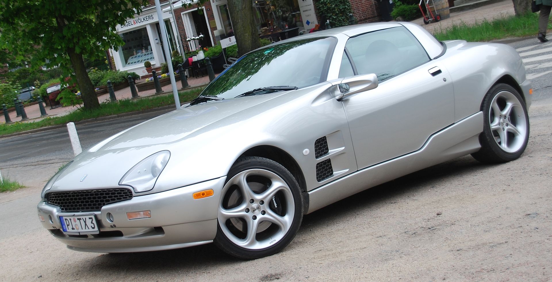Silver sports car (Qvale Mangusta) parked on urban street. Sleek convertible with distinctive front grille, stylized alloy wheels, positioned near trees