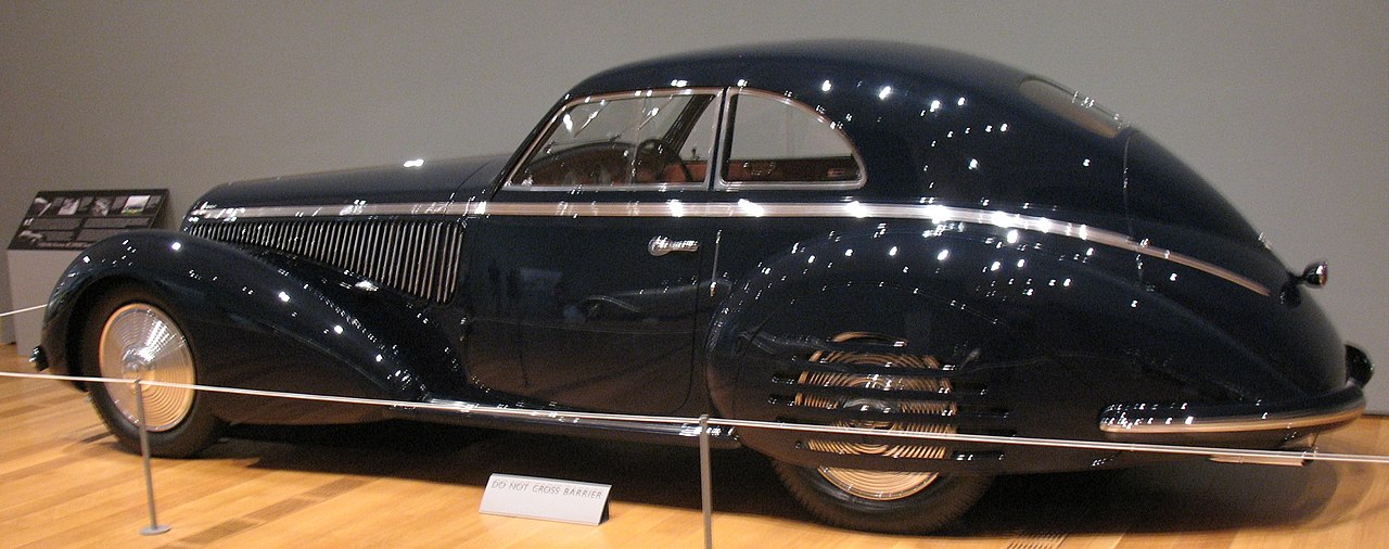 A sleek, black 1957 Alfa Romeo 8C 2900B Touring Berlinetta is displayed in a museum. The vintage car has an aerodynamic teardrop shape, chrome trim, and spoked wheels. A “Do Not Cross” sign and museum plaques are visible in the background
