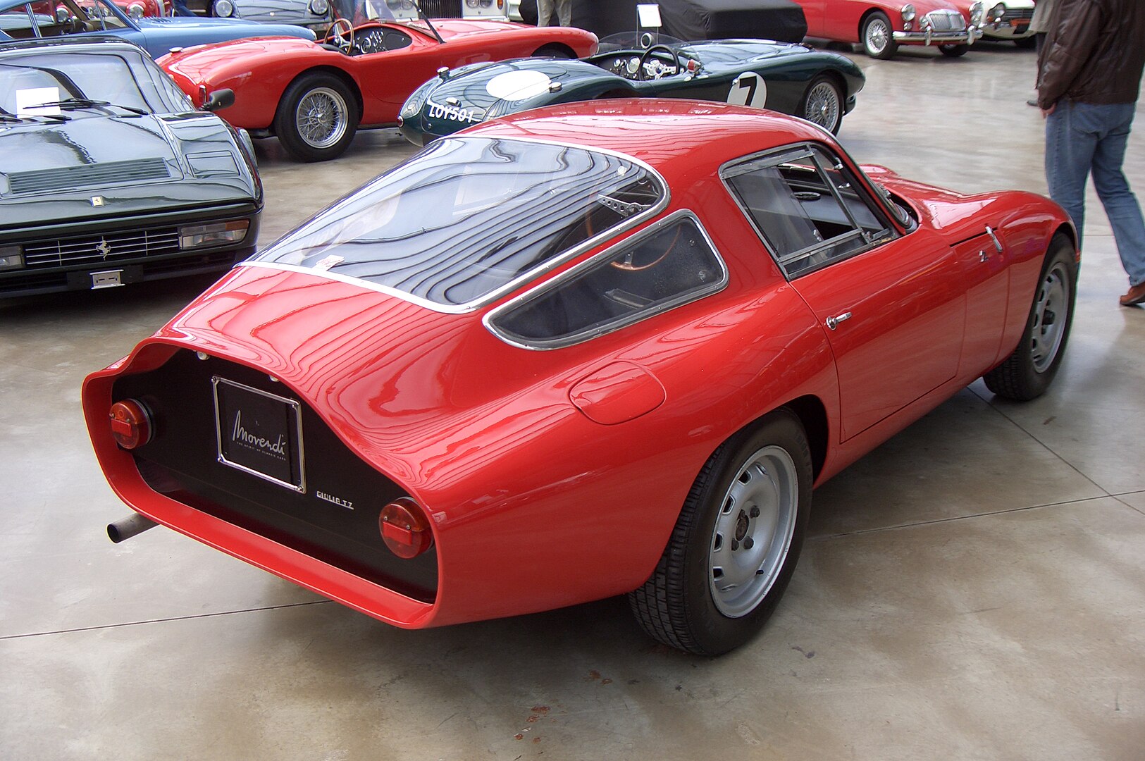 Bright red vintage Tubolare Zagato sports car with distinctive fastback design, displayed in indoor exhibition. Sleek aerodynamic body, curved rear window, multiple classic cars in background. Moverre branding visible