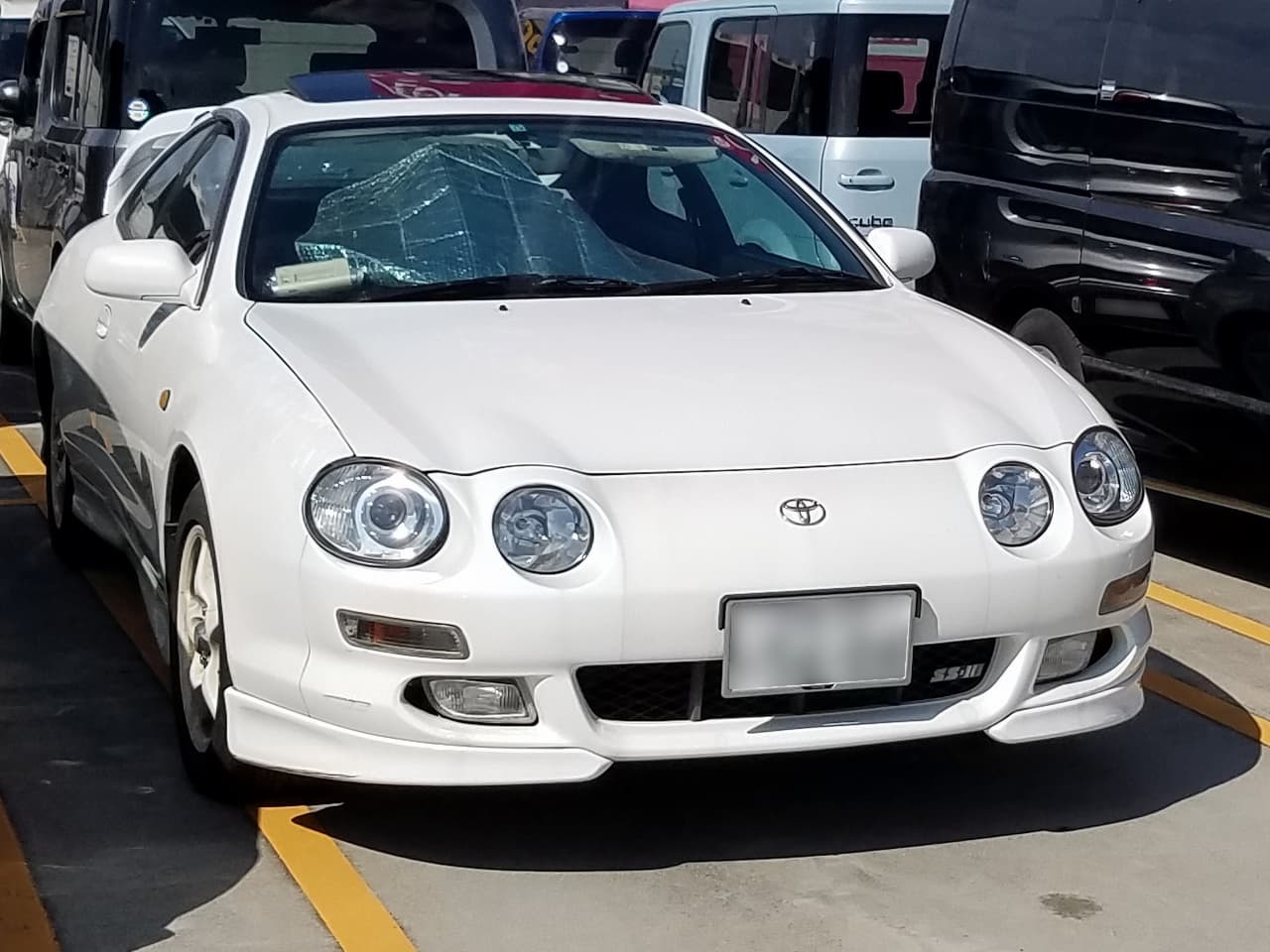 White Toyota Celica with distinctive round headlights parked in a parking lot, surrounded by other vehicles. Front view shows clean lines, sporty bumper, and aftermarket styling elements
