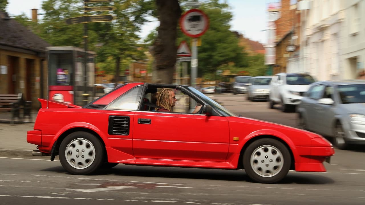 A red Toyota MR2 W10 with a targa top drives through a town. The car has a wedge-shaped design, side vents, and a rear spoiler. A man with dreadlocks is driving, while blurred cars, buildings, and traffic signs fill the background