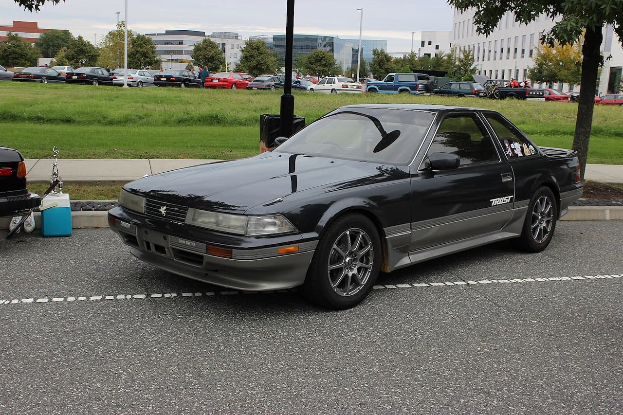 Black Toyota Crown or Toyota Crown Royal Saloon parked in a parking lot with "Trust" decal, featuring chrome wheels, against a backdrop of grass, trees, and office buildings