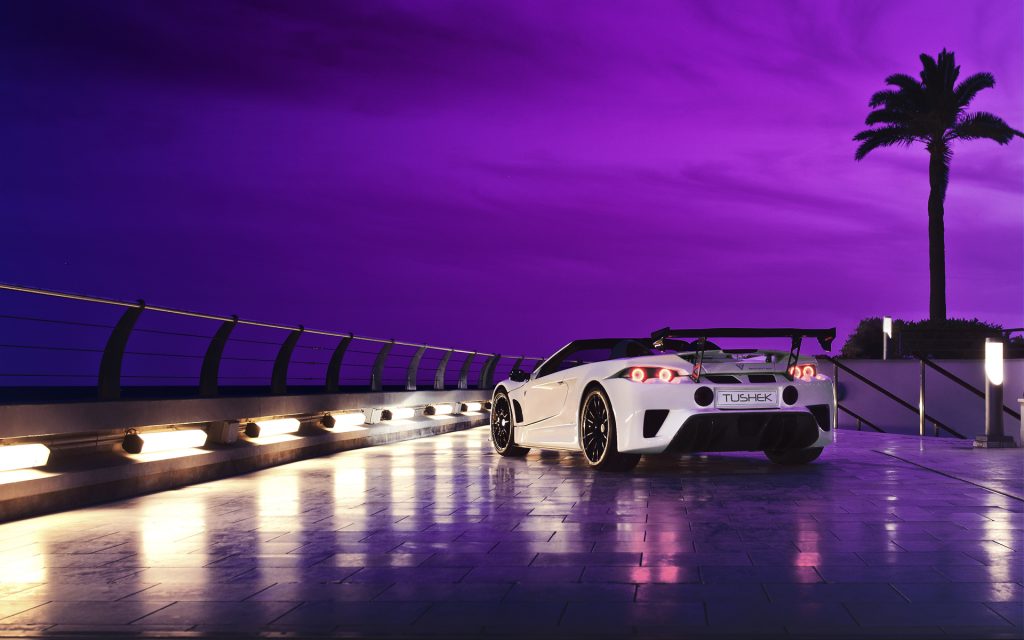 White sports car parked on illuminated platform at night, with dramatic purple sky and silhouetted palm tree. Sleek vehicle with rear spoiler, reflective taillights, against moody urban architectural backdrop