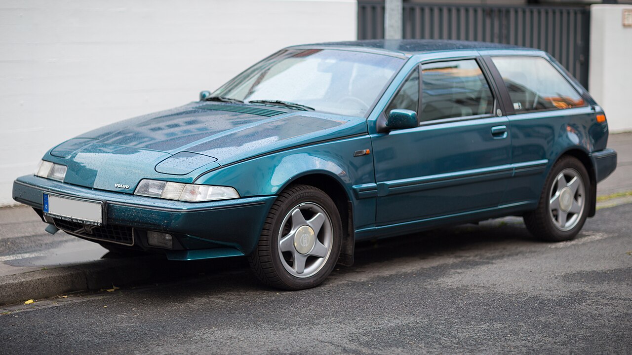 Teal-colored Volvo hatchback parked on asphalt street, side view. Metallic finish, five-spoke alloy wheels, clean lines characteristic of late 20th-century automotive design. Background features urban setting with metal fence