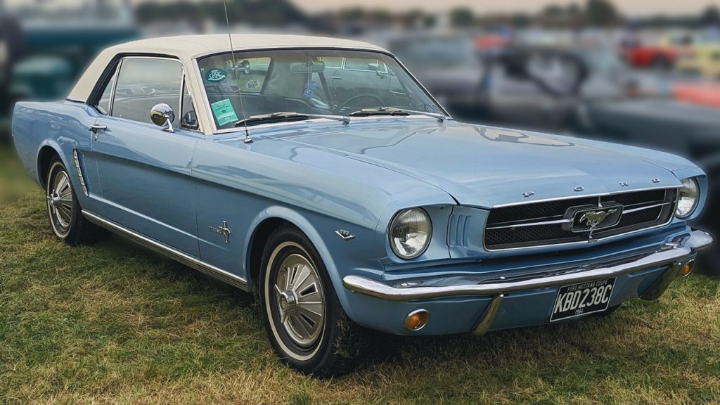 Classic light blue 1964 Ford Mustang convertible parked on grass with chrome details and white soft top at car show