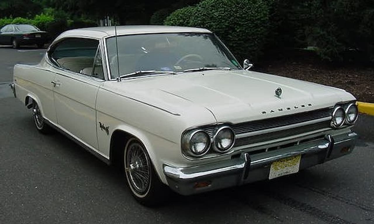 A classic white 1965 AMC Marlin coupe with a sleek fastback roofline is parked on a paved surface. It features quad headlights, a chrome grille, and a long hood. Wire-spoke wheels and chrome bumpers enhance its vintage appeal, with trees and another car in the background
