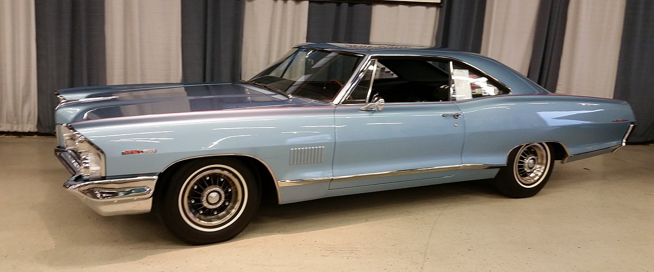 A classic light blue 1965 Pontiac 2+2 coupe with chrome trim, black wheels, and a sleek, elongated body, displayed indoors against a backdrop of white and dark curtains. The car's polished surface reflects the showroom lights