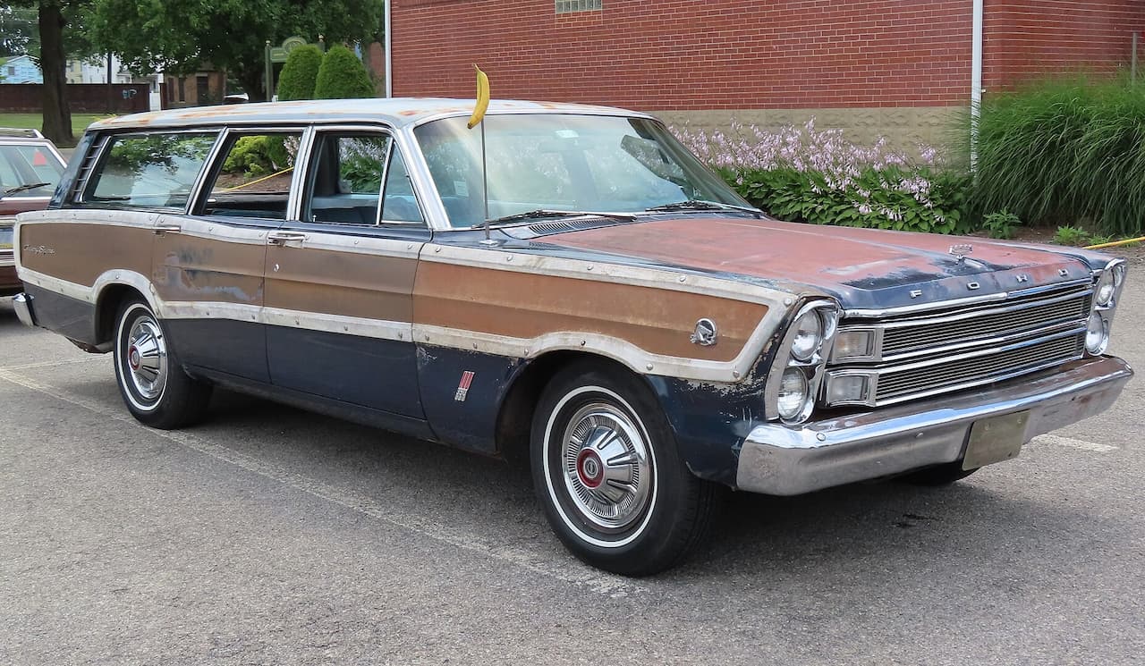 A weathered 1966 Ford Country Squire station wagon with faded wood paneling and worn blue paint is parked on pavement. It features chrome trim, round hubcaps, and a long body. A banana-shaped antenna topper stands out. A brick building, greenery, and another vintage car are in the background