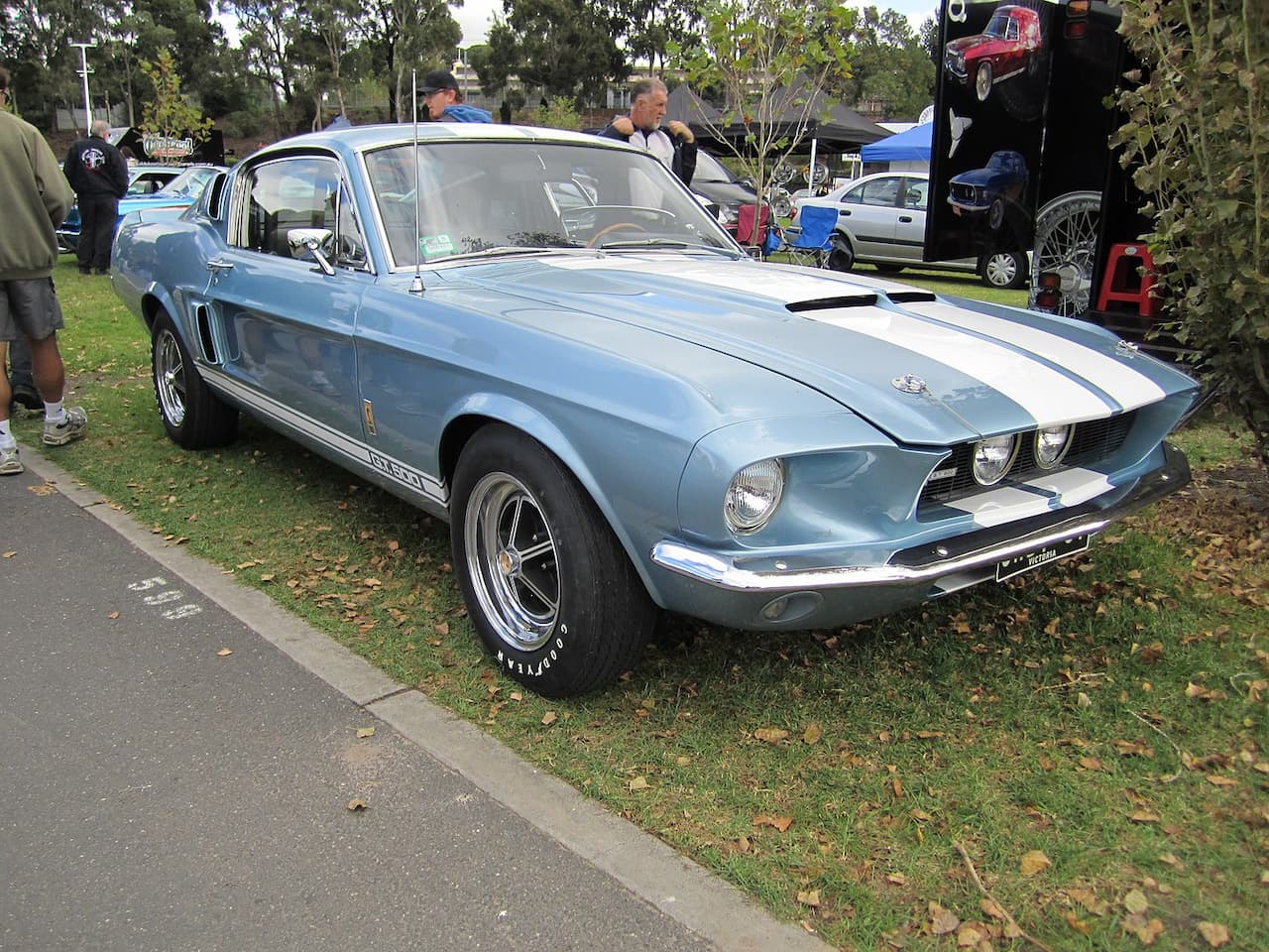 A light blue 1967 Shelby GT500 Mustang with white racing stripes is parked on grass at an outdoor car show. It features chrome wheels, Goodyear tires, hood pins, and a Cobra badge