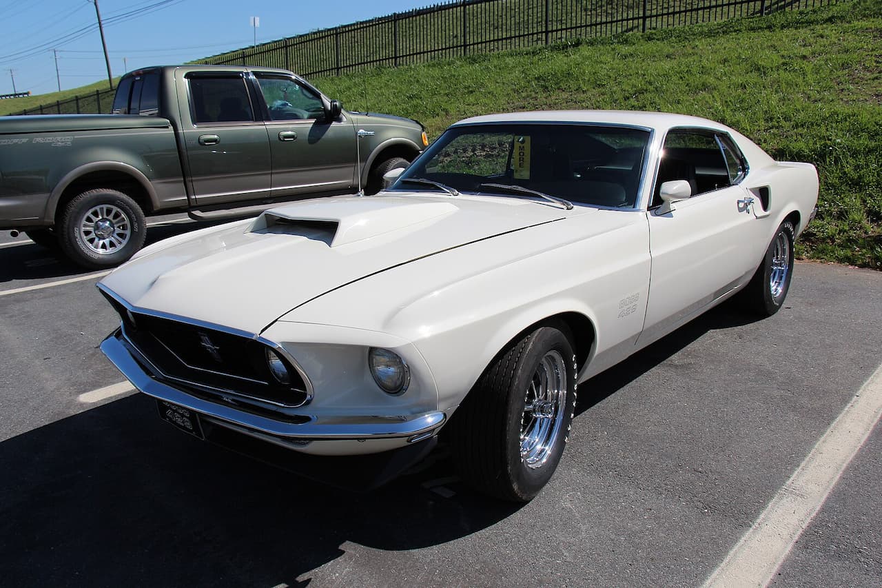 A classic white 1969 Ford Mustang Boss 429 is parked in an outdoor lot. The car features a blacked-out grille, chrome bumpers, a large hood scoop, and polished wheels. A green Ford F-150 pickup truck is parked behind it near a grassy hill