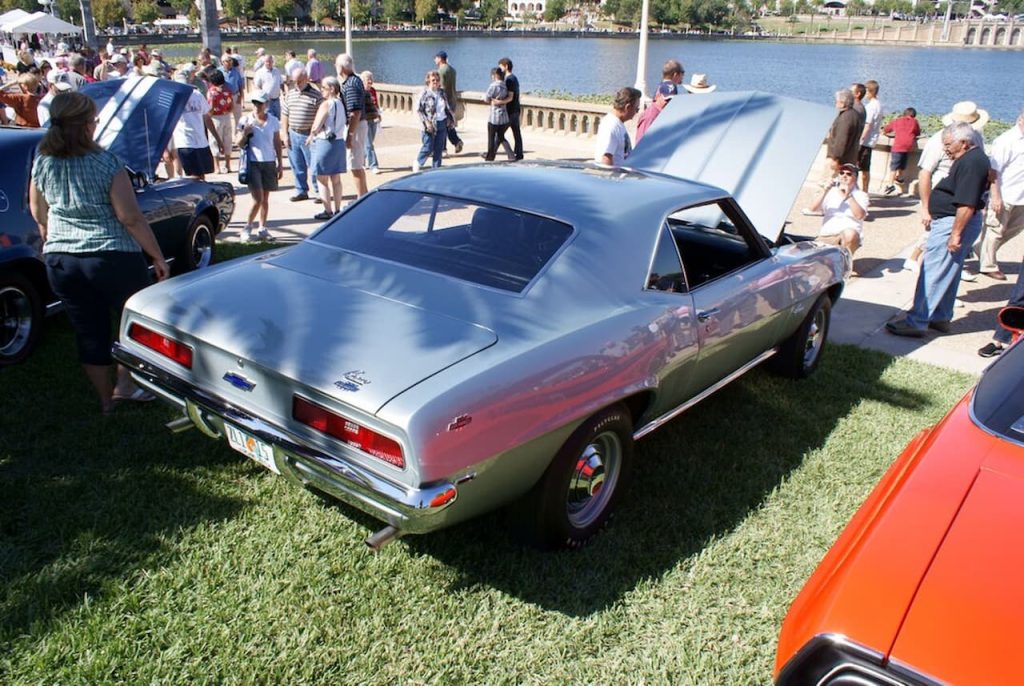 A silver 1969 Chevrolet Camaro ZL1 is displayed at a car show near a waterfront. The car has a sleek body, chrome bumpers, and red taillights. A crowd of attendees, other classic cars, and a scenic lake with trees are visible in the background