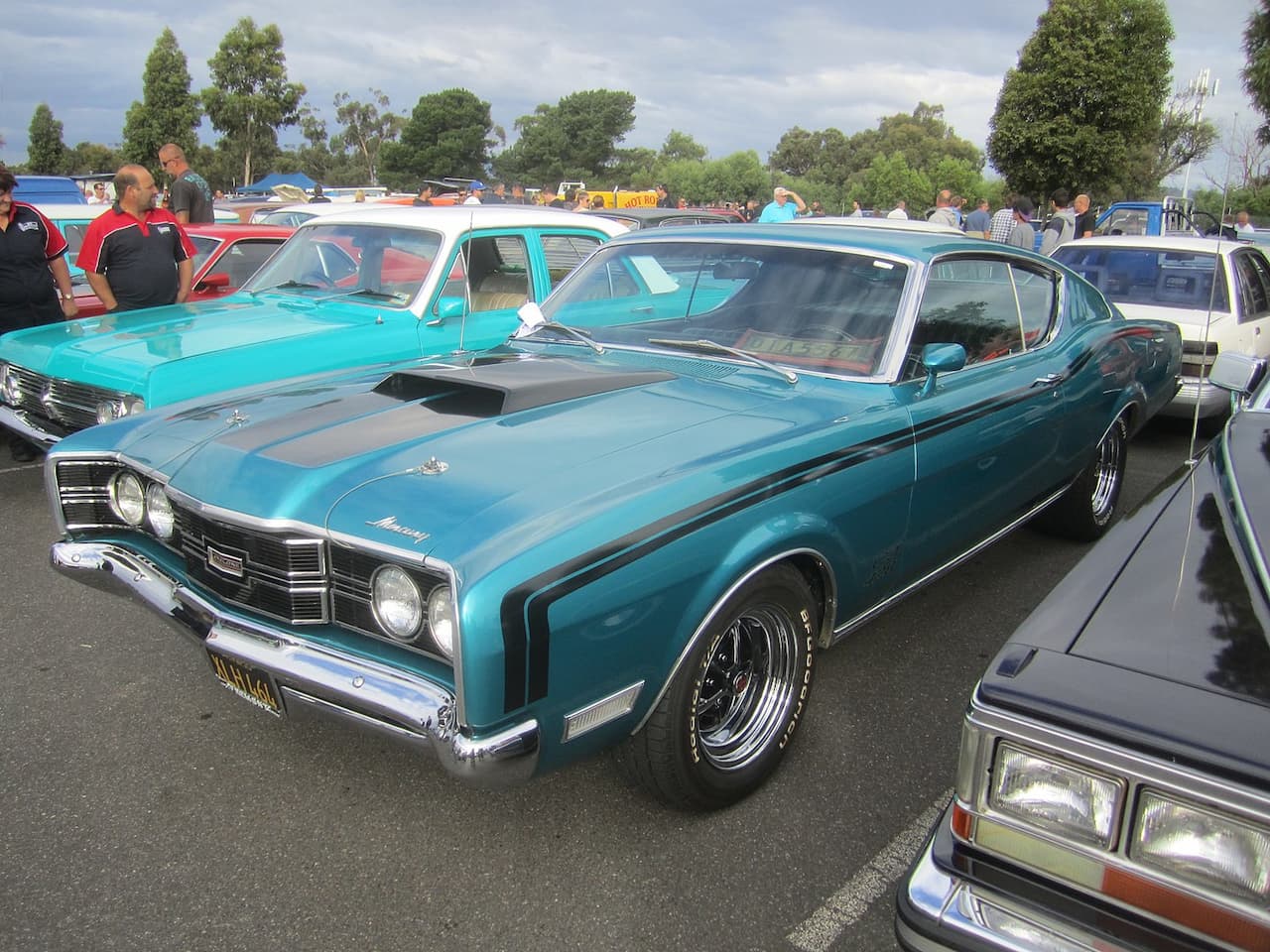 A classic blue 1969 Mercury Cyclone CJ with black racing stripes and a hood scoop is parked at a car show among other vintage vehicles. Chrome bumpers, polished wheels, and a crowd of people admiring the cars are visible under a cloudy sky