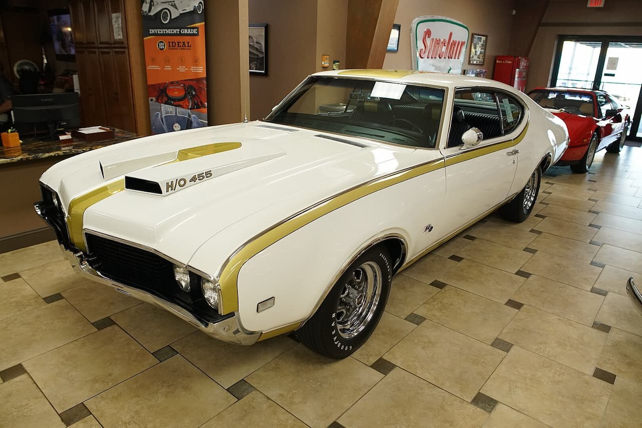 Classic white and gold Oldsmobile Hurst/Olds muscle car with H/O 455 badging. Displayed in a showroom with tiled floor, vintage automotive posters, and Sinclair sign. Chrome wheels, sporty design characteristic of late 1960s American muscle cars