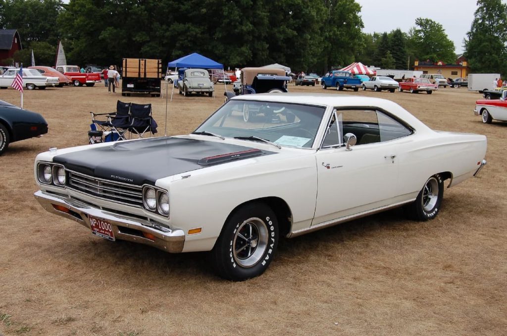 Classic white and black Plymouth muscle car at a vintage car show. Parked on a grassy field with other classic vehicles, tents, and flags in the background, showcasing a nostalgic automotive event