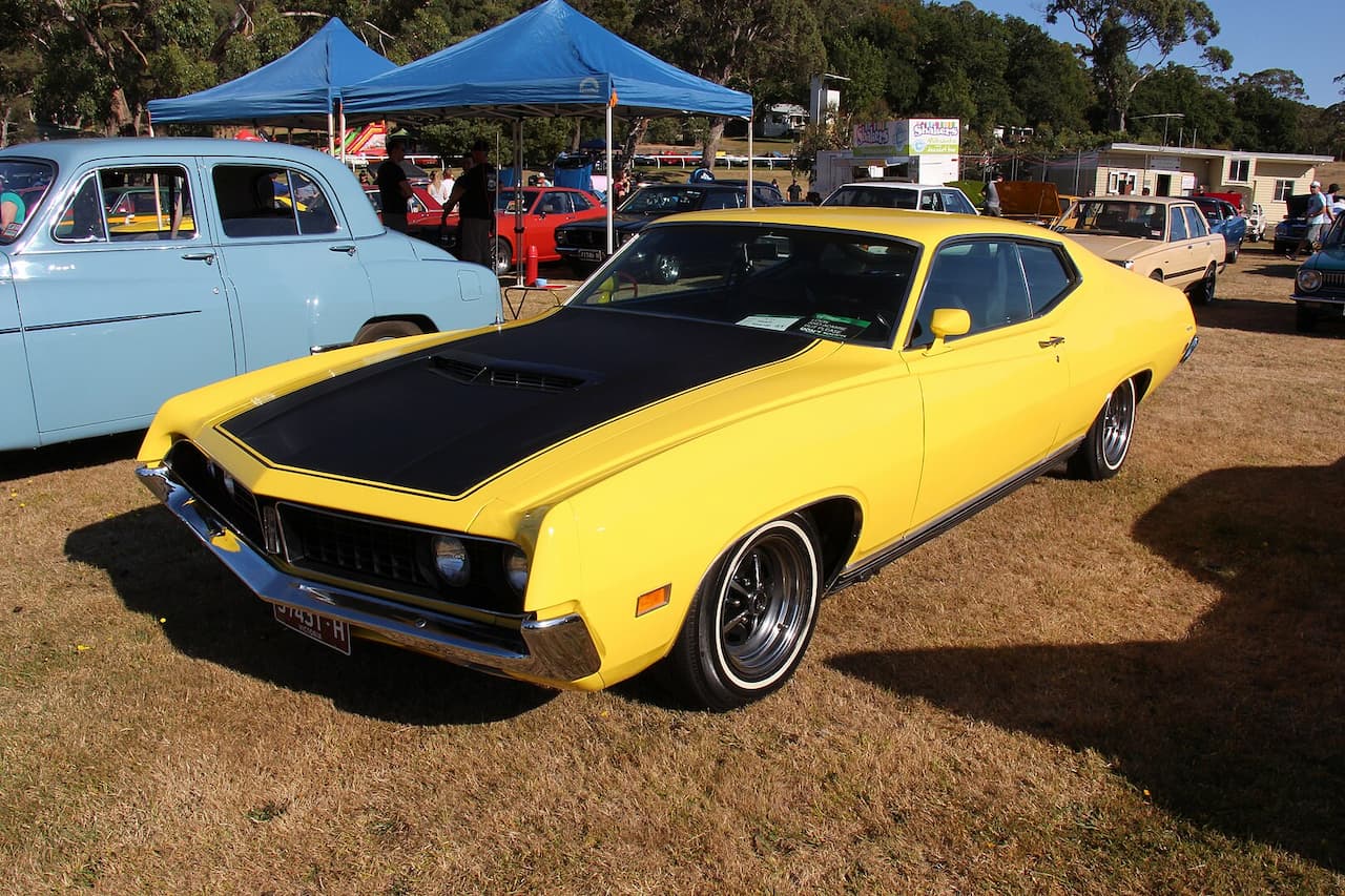 Bright yellow muscle car with black hood at a vintage car show. Parked on dry grass with blue canopy tents and other classic vehicles in the background. Chrome details gleam in sunlight