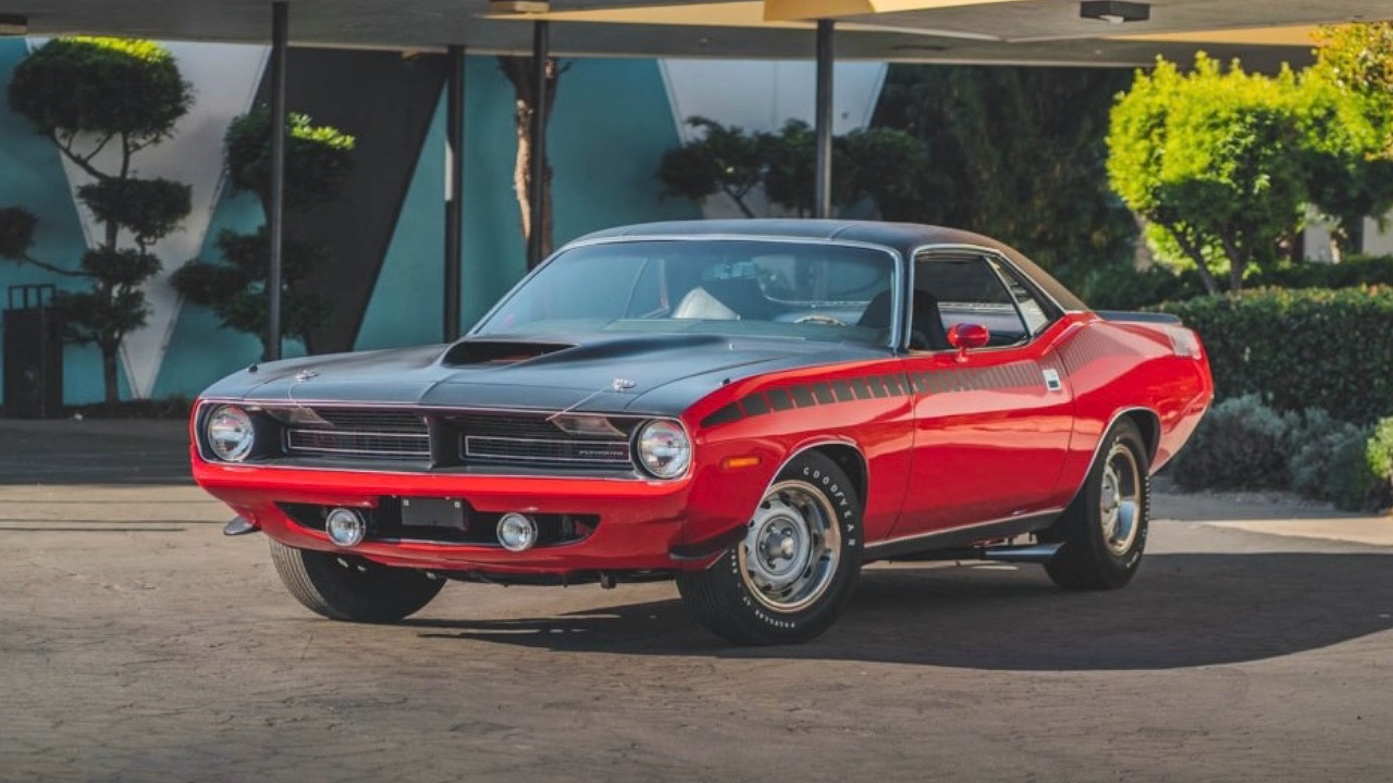 Bright red vintage muscle car, likely a Plymouth Barracuda, parked under architectural overhang. Sleek automotive design with chrome details and performance-oriented styling, showcasing classic American muscle car aesthetics