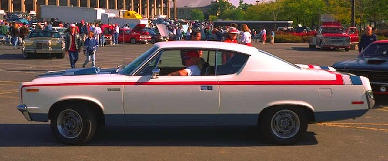 A white 1970 AMC Rebel Machine with red, white, and blue stripes is parked at a car show. It has a fastback roofline, chrome bumpers, and rally wheels. A crowd, classic cars, and a stadium are visible in the background under bright sunlight