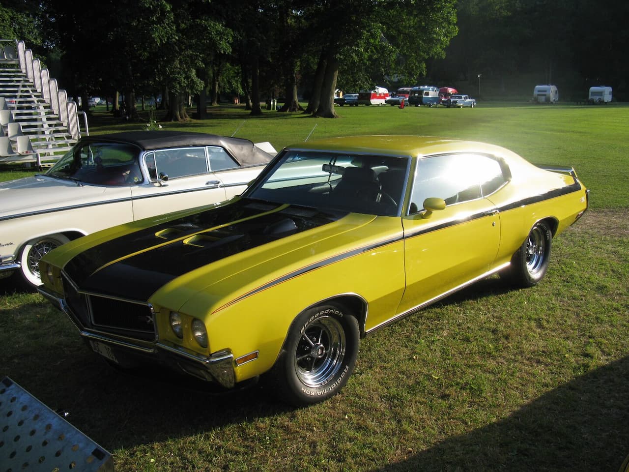 Yellow and black Buick muscle car with distinctive racing stripes, parked on green grass at a vintage car show. Trees and other classic vehicles visible in the background under soft sunlight