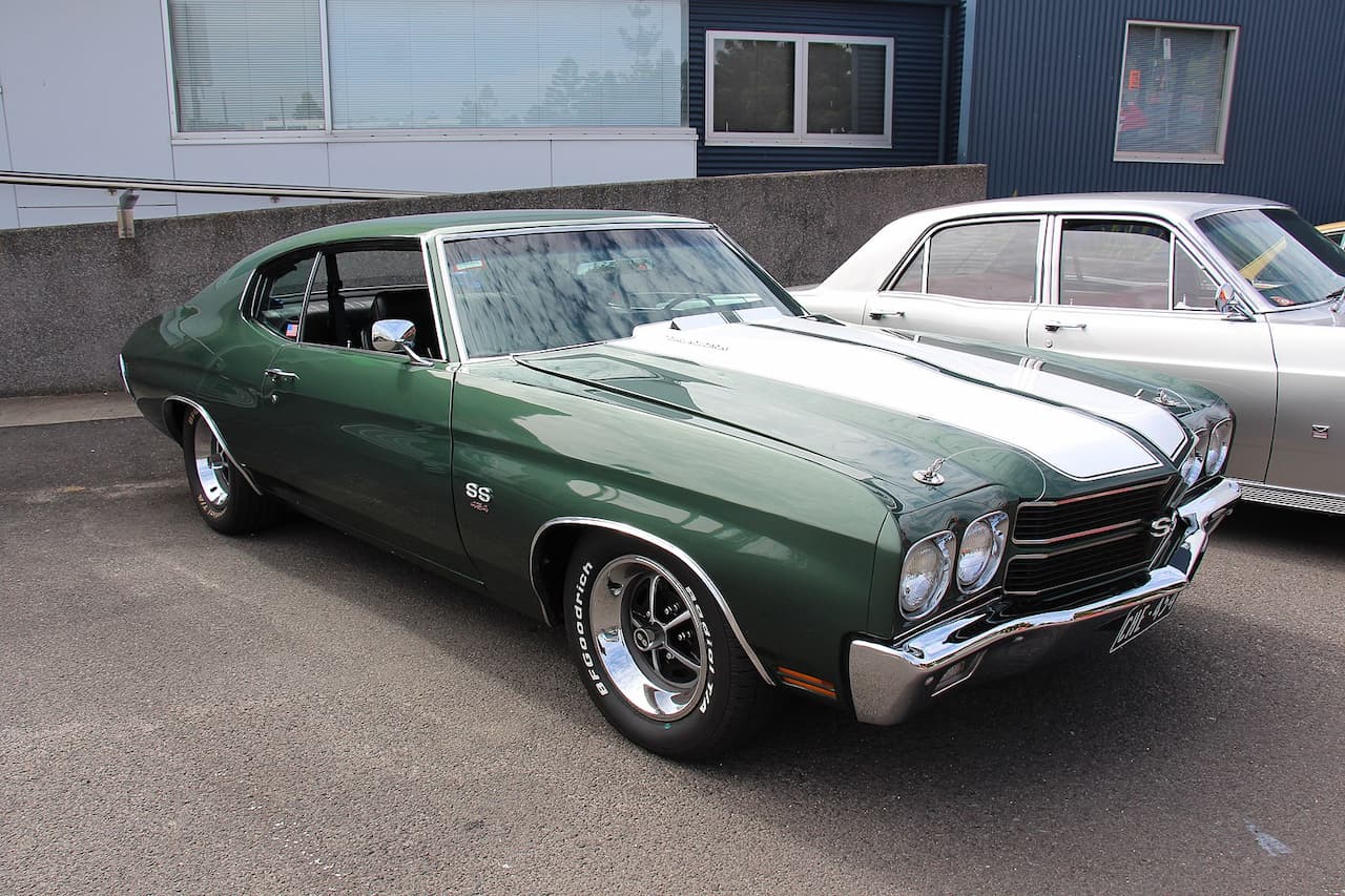 A classic 1970 Chevrolet Chevelle SS 454 in dark green with white racing stripes, parked outdoors. The muscle car features chrome bumpers, SS badging, BF Goodrich tires, and a black interior. A vintage white sedan is partially visible behind it