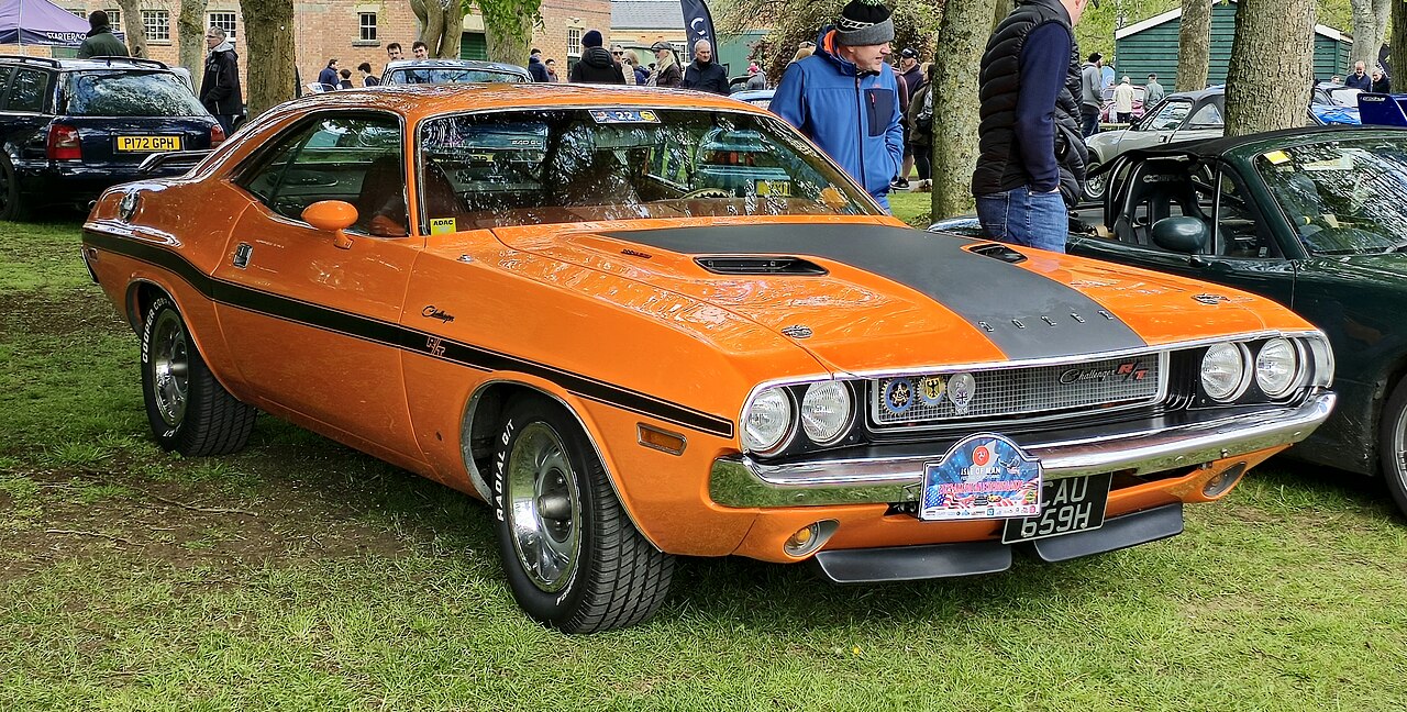 A vibrant orange 1970 Dodge Challenger R/T with black racing stripes is displayed at a car show. The muscle car features chrome bumpers, a black grille, and rally wheels. A crowd of attendees and classic cars are visible in the background under trees