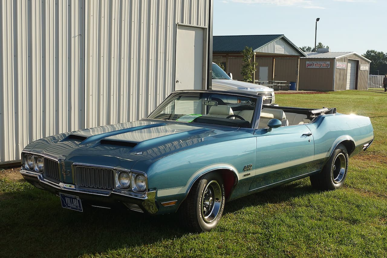 A light blue 1970 Oldsmobile 442 W-30 convertible with white interior sits on a grassy field near a metal-sided building. It features a black soft top, dual hood scoops, chrome bumpers, and rally wheels. A silver SUV and sheds are visible in the background