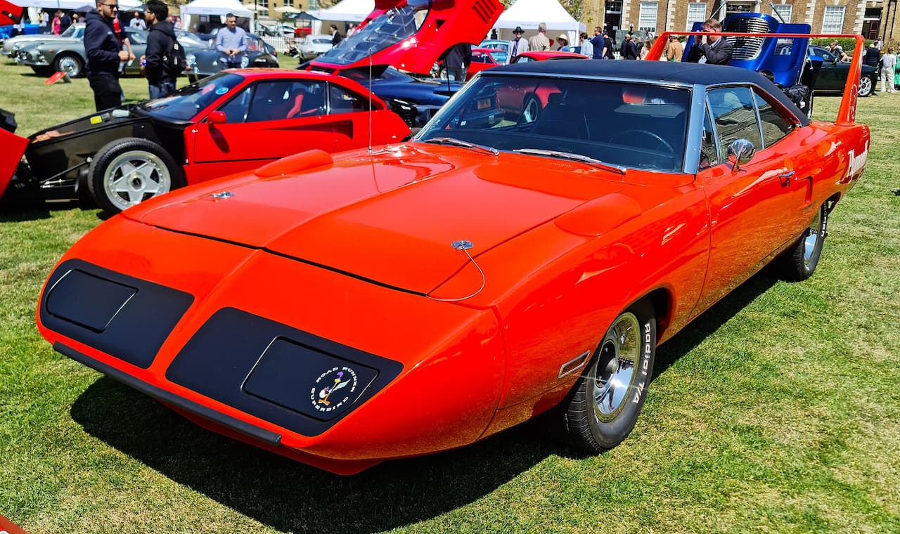 A bright orange 1970 Plymouth Road Runner Superbird with a black vinyl top is displayed at an outdoor car show. It features a long aerodynamic nose, pop-up headlights, a massive rear wing, and Road Runner badging. Other classic and exotic cars, along with people, surround it