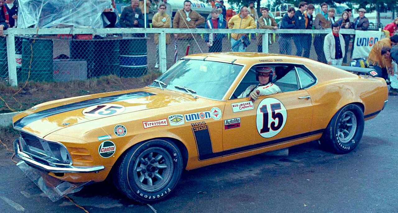 Yellow vintage racing car with number 15, marked with sponsor logos. Appears to be a classic sports car positioned at a racing event or exhibition, showcasing motorsport heritage