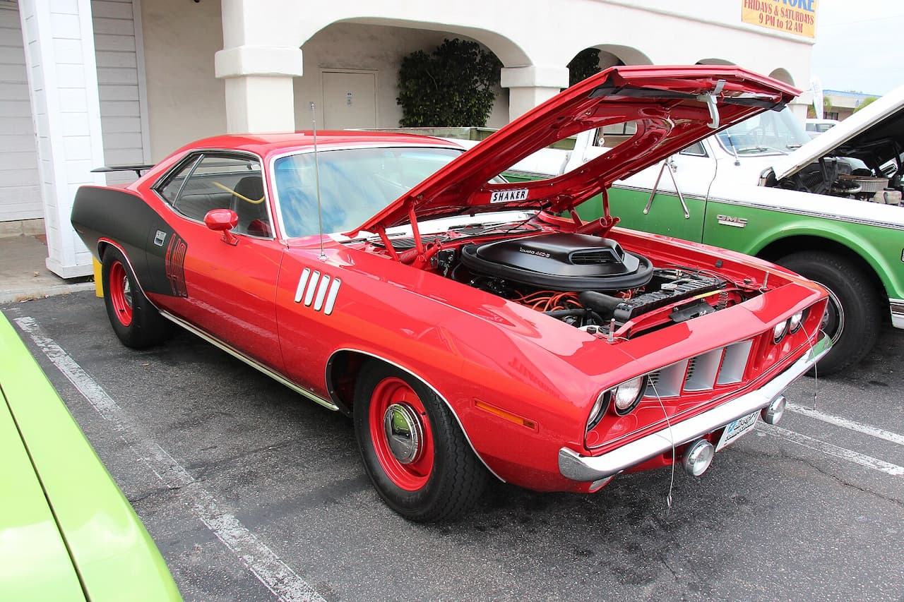 A red 1971 Plymouth HEMI 'Cuda with a black rear section and red steel wheels is displayed at a car show. The hood is open, revealing a powerful 426 HEMI engine with a Shaker scoop. Other classic cars and a white building are in the background