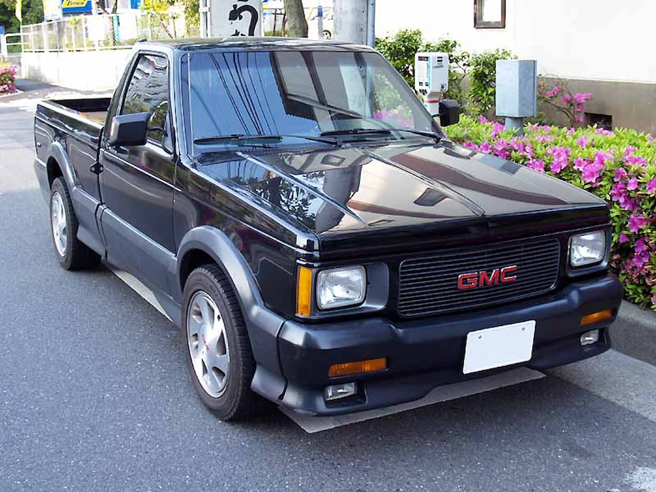 A black 1992 GMC Syclone pickup truck parked on a street. It has a sleek body, red GMC badging on the grille, flared wheel arches, and silver alloy wheels. The background features pink flowers, a sidewalk, utility poles, and buildings