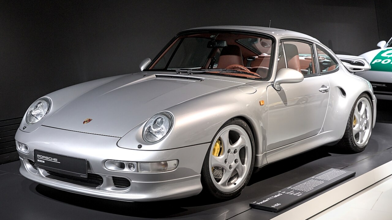 Silver Porsche sports car, side view. Sleek curves, distinctive rounded headlights, yellow-rimmed alloy wheel. High-performance design captured in studio setting with dramatic lighting