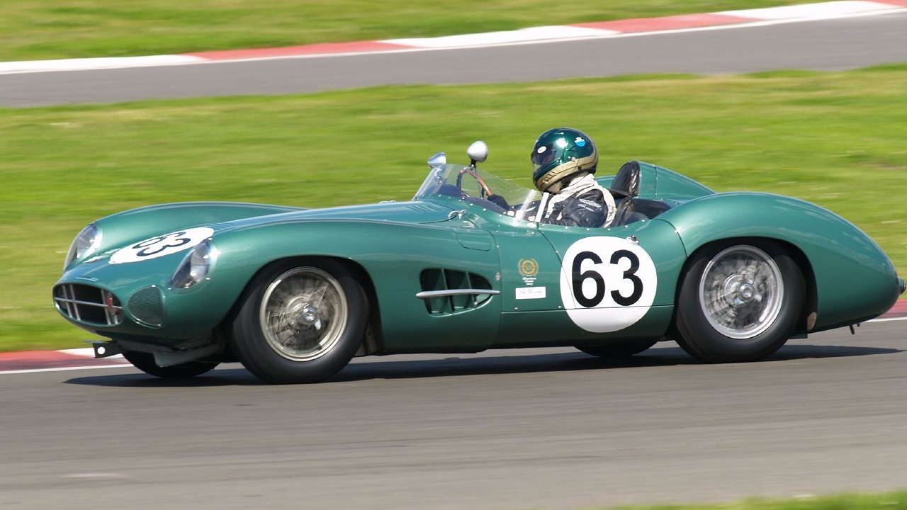 A vintage green race car with the number 63 speeds on a racetrack. The open-top vehicle features aerodynamic curves, a low profile, and a side vent. The driver, wearing a blue helmet and racing suit, is gripping the steering wheel