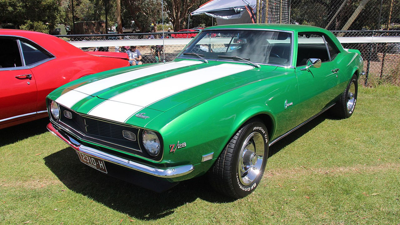 Vintage green muscle car with white racing stripes, parked on grass. Classic American sports car design with chrome details, showcasing a pristine restoration or well-maintained condition at what appears to be a car show or exhibition