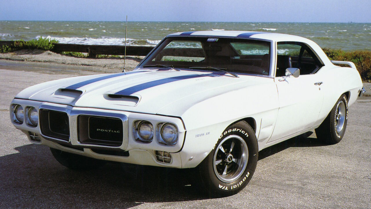 Classic white muscle car with blue racing stripes, featuring a distinctive front grille and powerful stance. Photographed in a parking lot or display area with a clear sky in the background