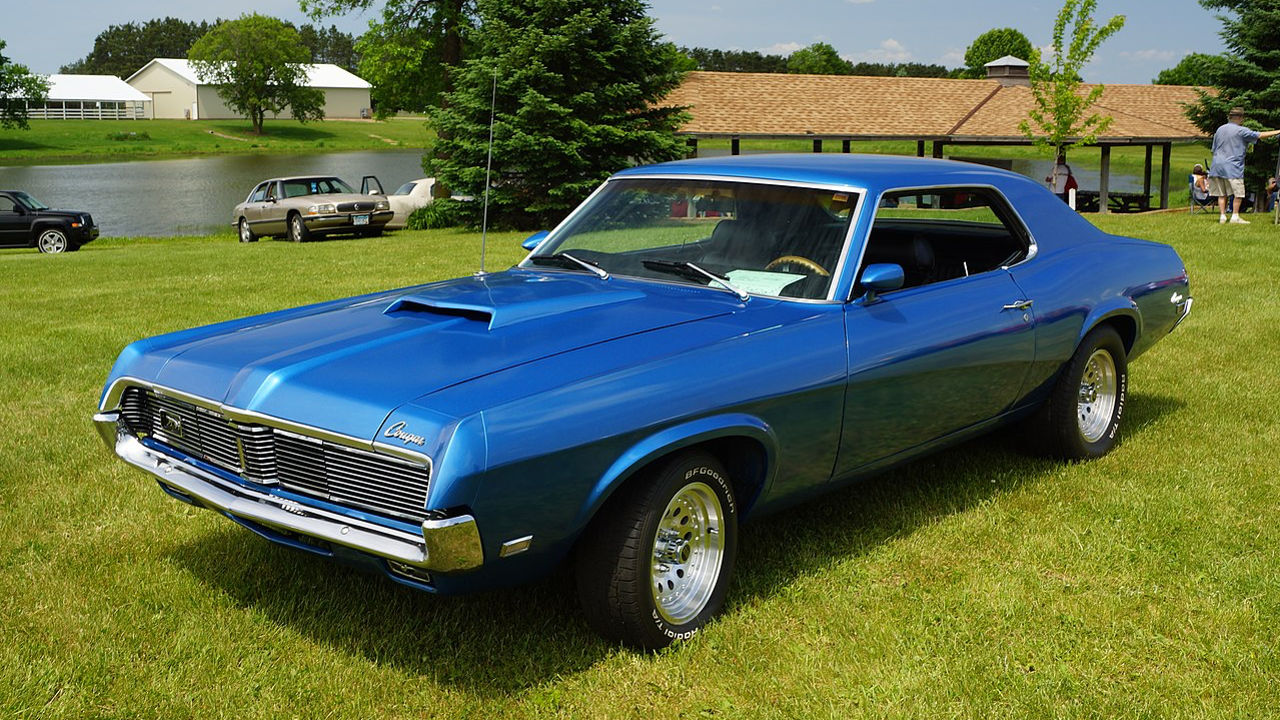 Classic blue Mercury Cougar muscle car parked on green grass. Sleek automotive design with chrome details, hood scoop, and polished wheels. Background features a white building and trees