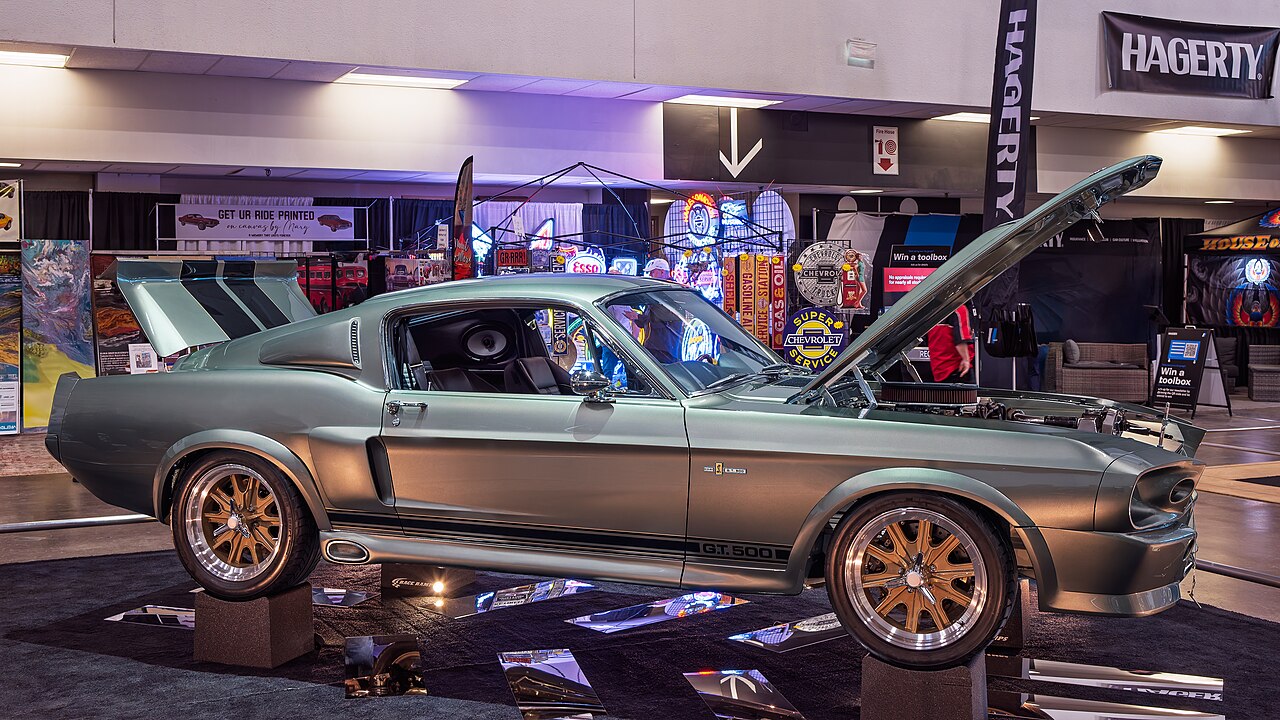 Silver 1967 Shelby GT500 muscle car with hood open displayed at automotive show with vintage signage backdrop