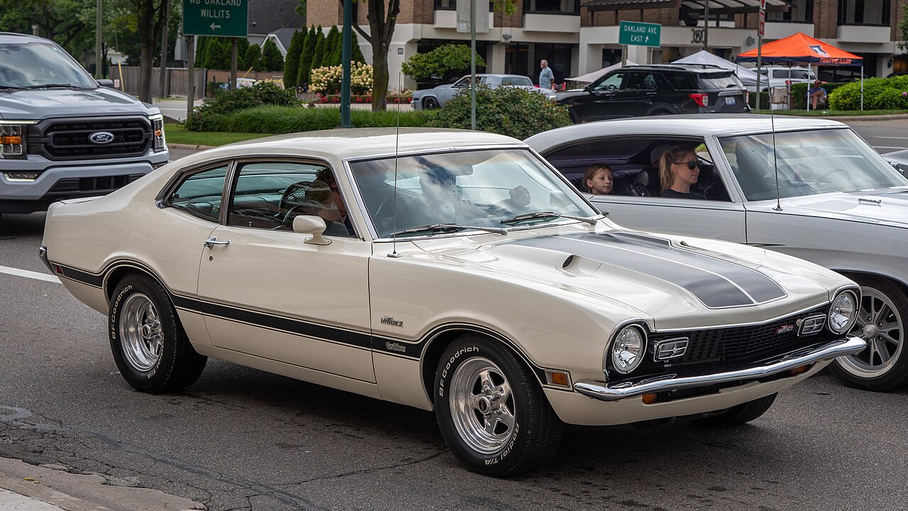 White 1971 Ford Maverick Grabber classic muscle car with racing stripes parked on street at car show event