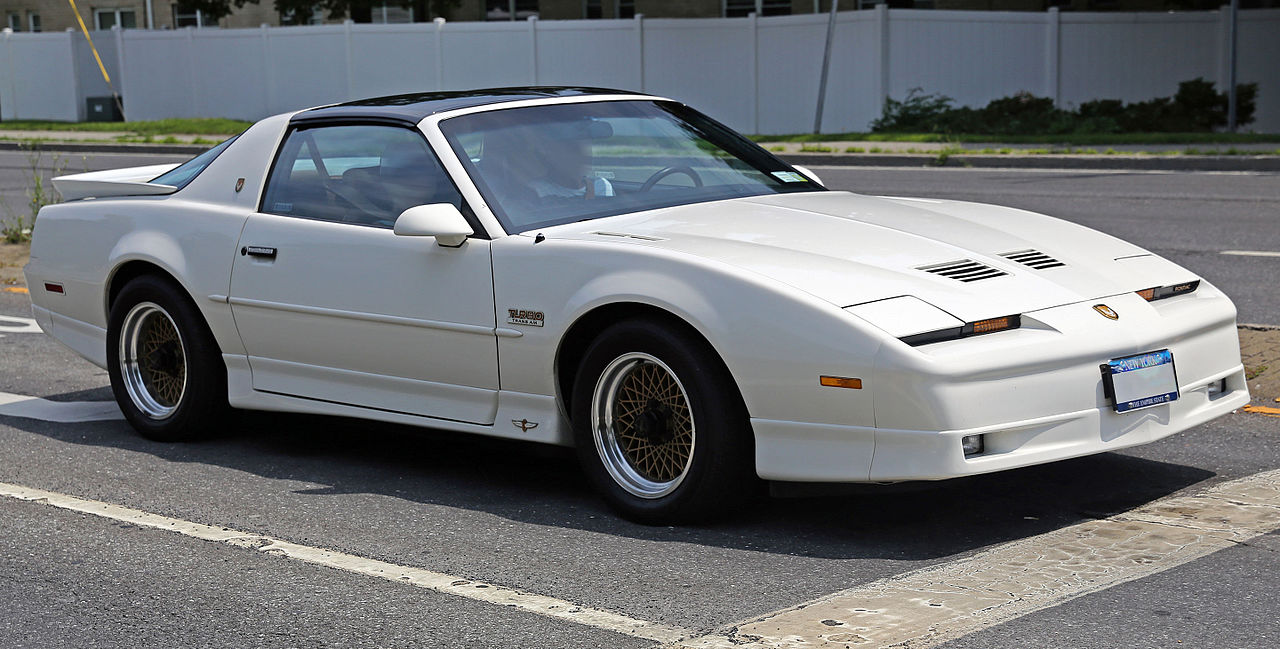 White 1989 Pontiac Firebird GTA with T-tops and distinctive wheels parked in parking lot near white fence