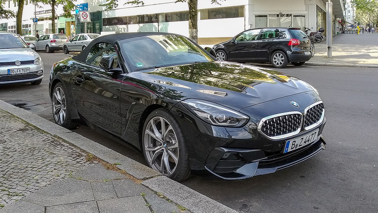 Black 2025 BMW Z4 convertible sports car parked on European street with cobblestone sidewalk and other vehicles nearby