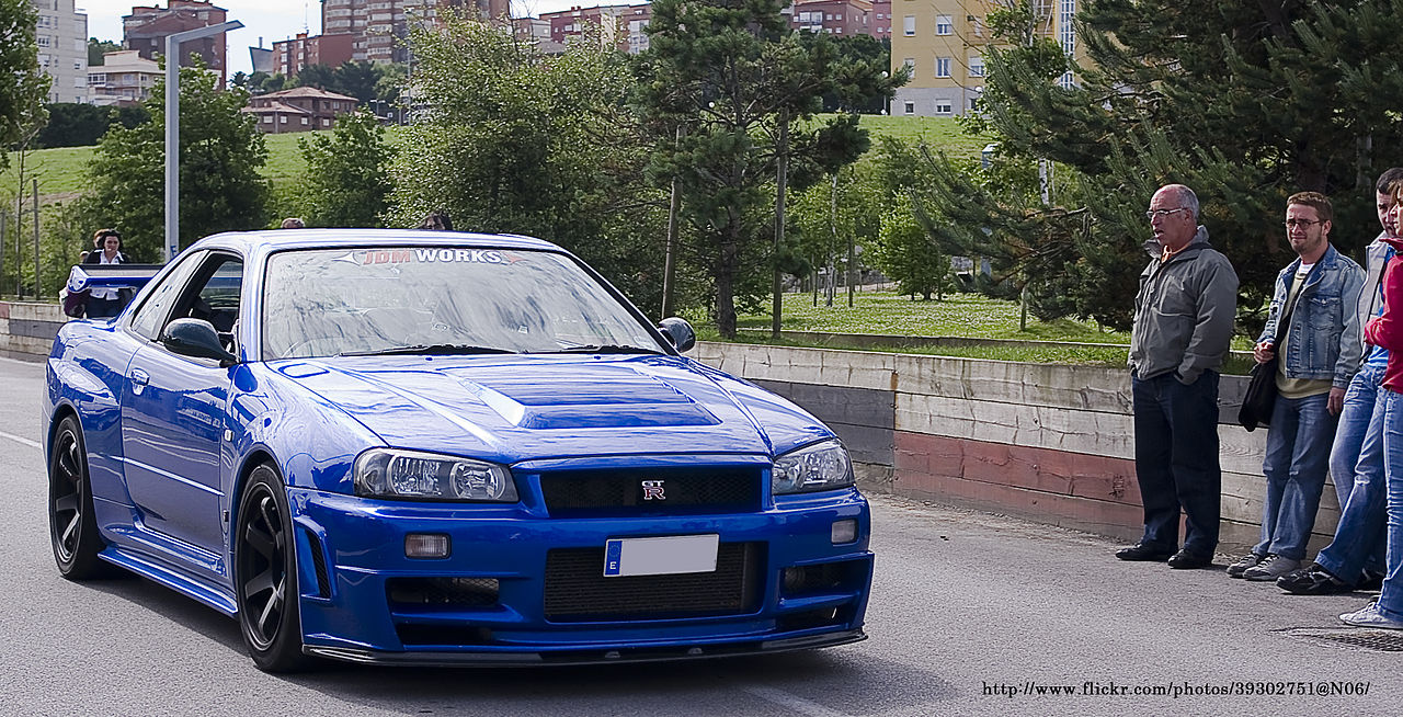 Blue Nissan Skyline GT-R R34 with aggressive body kit and black wheels at car meet with spectators