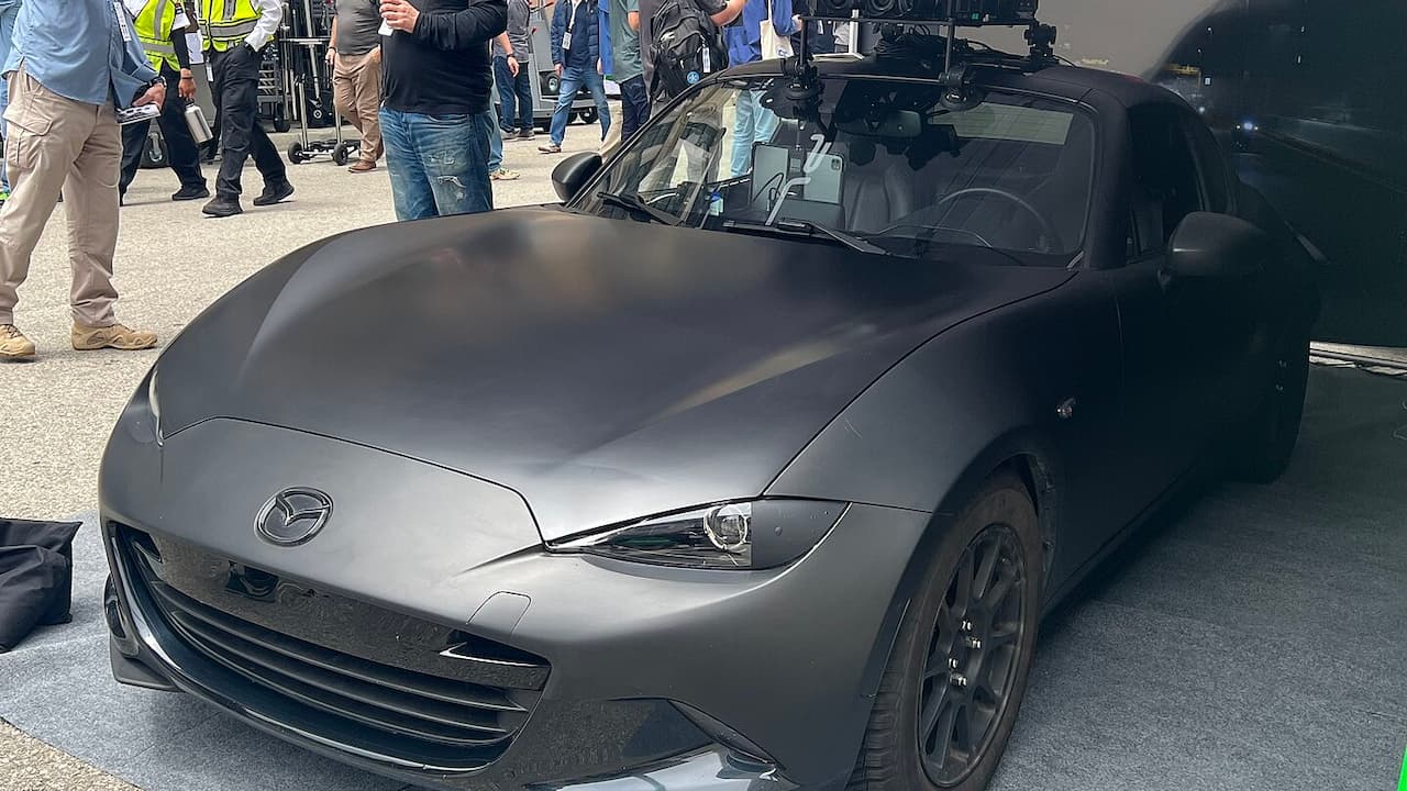 Gray 2025 Mazda MX-5 Miata with soft top down displayed at automotive event with crowd and camera equipment nearby