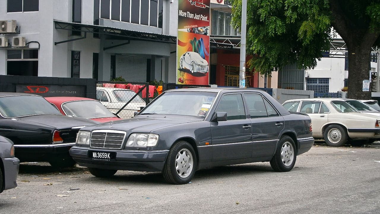 Gray Mercedes-Benz E-Class (W124) parked in urban lot with Malaysian license plate among other vintage cars and buildings