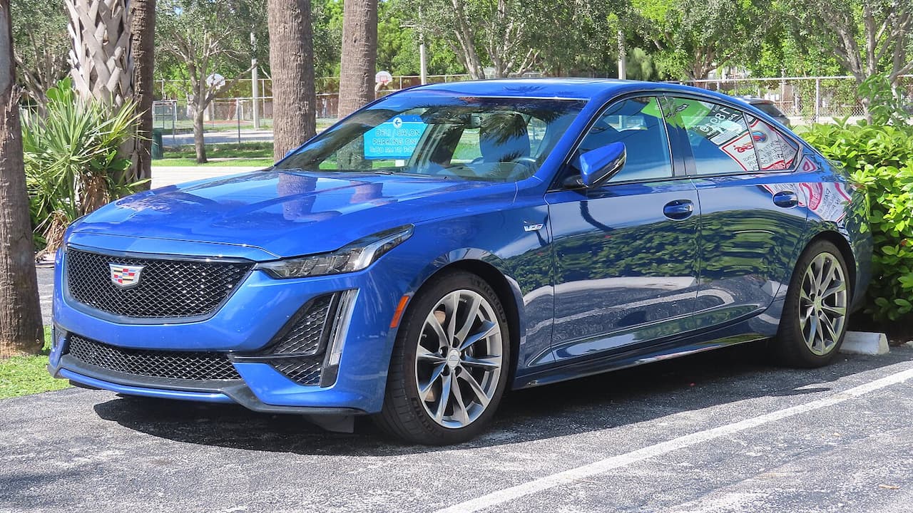Blue Cadillac CT5-V parked on street with distinctive mesh grille, large alloy wheels, and aggressive styling in tropical setting