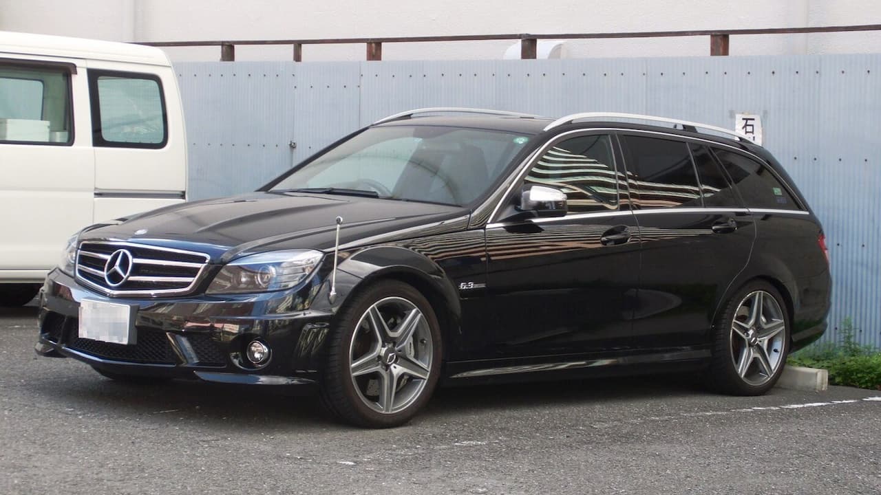 Black Mercedes-AMG C63 S wagon parked on asphalt with distinctive grille, AMG wheels, and white van in background