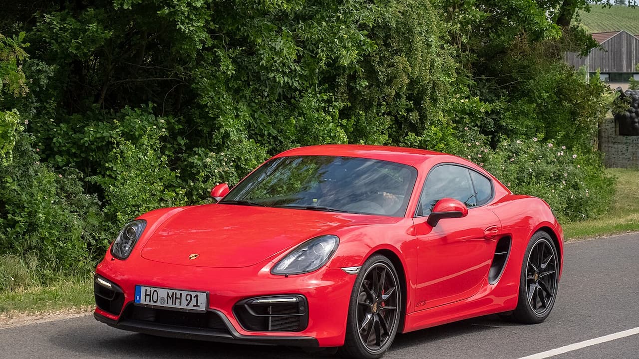 Red Porsche Cayman sports car parked on rural road with lush green trees and countryside buildings in background