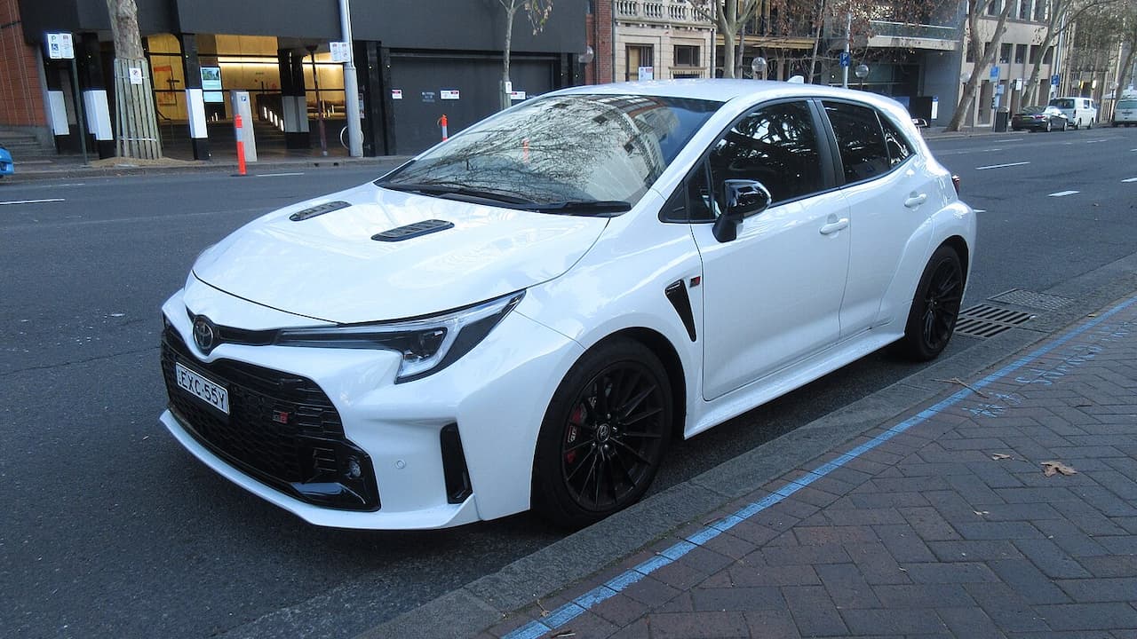 White Toyota GR Corolla hot hatch with black wheels, hood vents, and aggressive body kit parked on European city street