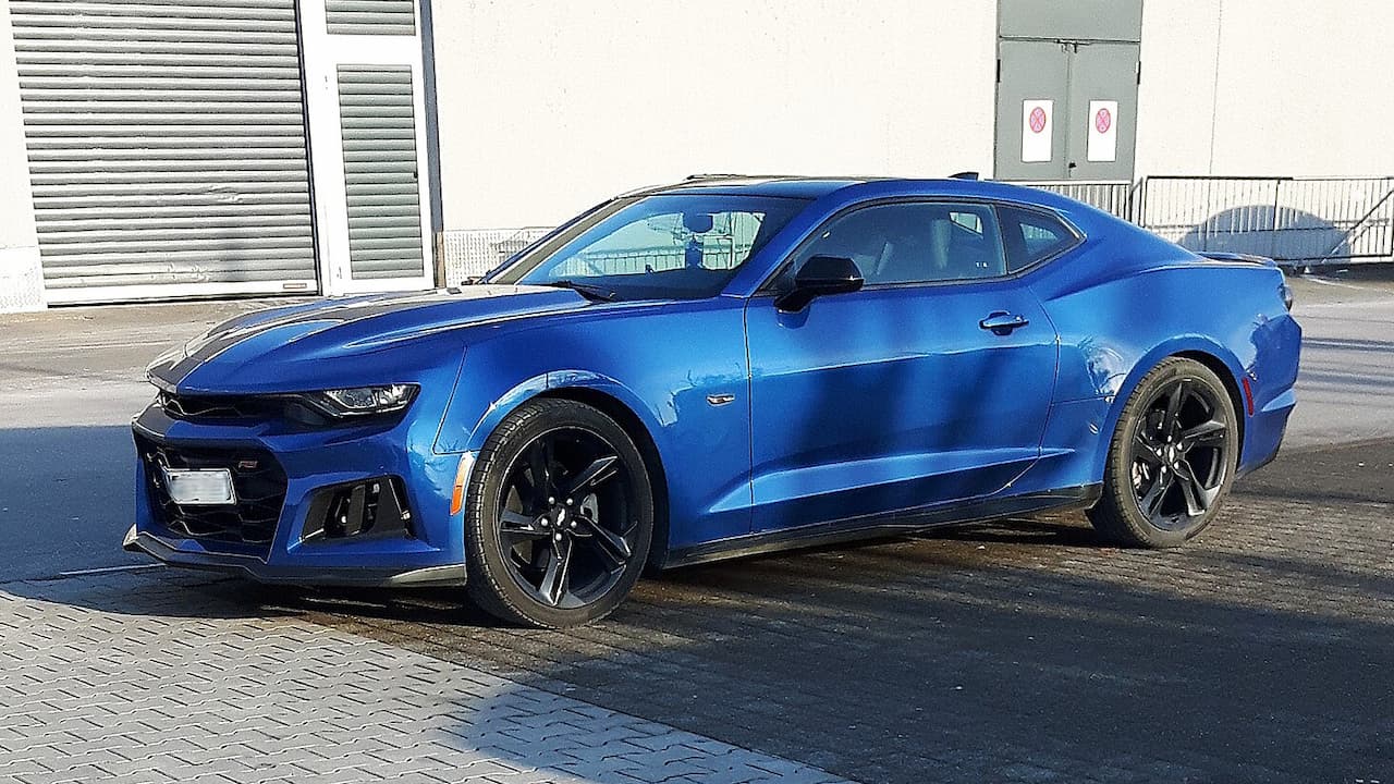 Blue 2025 Chevrolet Camaro sports car with black racing stripes parked on brick pavement in front of industrial building