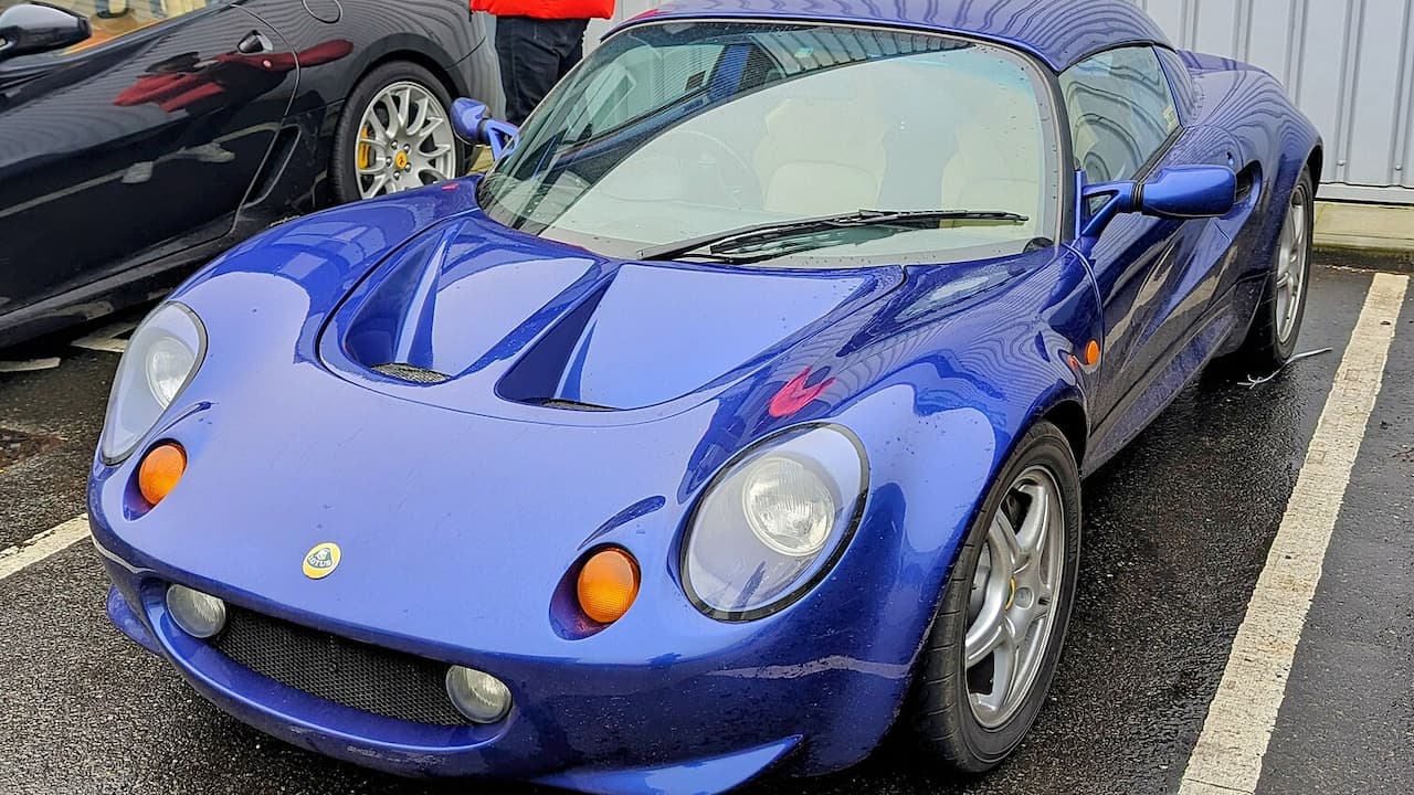 Blue Lotus Elise S1 sports car parked in lot with round headlights, orange side markers, and distinctive low-profile design